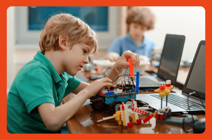 At a table, a young boy tinkers with a handmade mechanical object made with Legos and wires using a small screw.