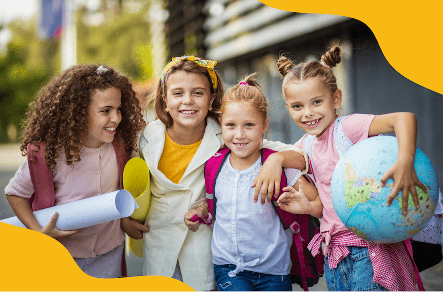 Four students smiling in front of a school. Two of the young girls carry rolled up posters while one of the far right holds a globe.