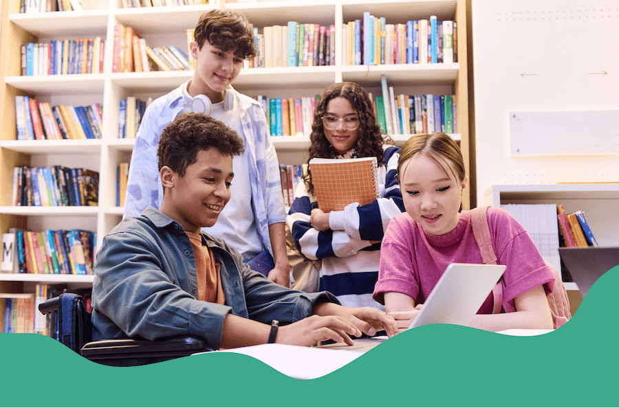 In a library, four students are gathered in the book stacks. A young boy and girl sit at a table working on a laptop with two more students standing behind them.