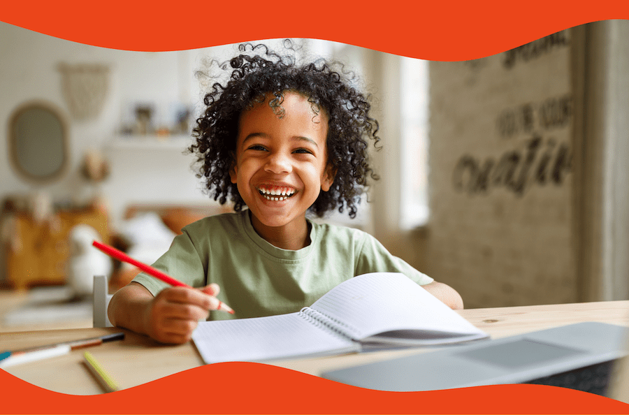 A smiling young boy sits at a desk with a red colored pencil. On the table are different colored pencils, an open notebook, and a laptop.