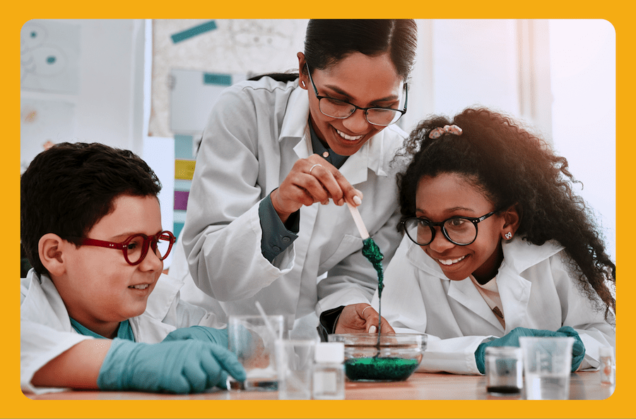In a science classroom, two students watch their teacher, a young woman experimenting with a green gooey substance in a bowl, all in lab coats.