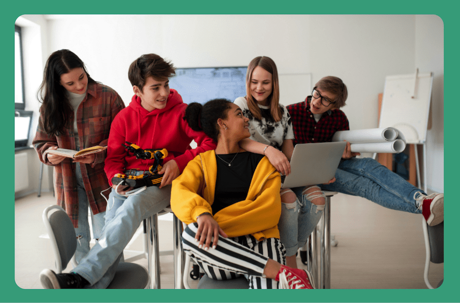 Five students sit together in an open classroom, holding different objects: a book, a robotic arm, a laptop, and two rolls of drawing paper.