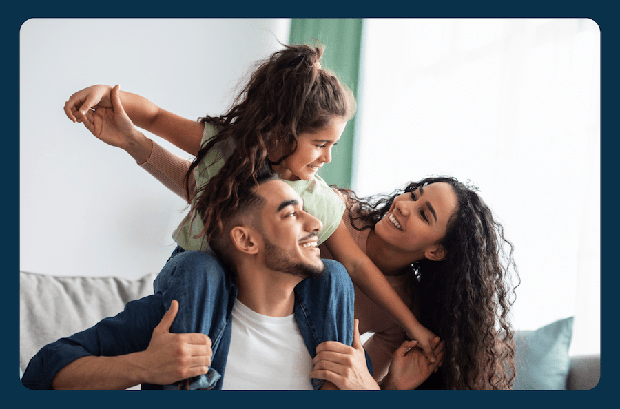 A family of three sit in a brightly lit living room. The father holds his young daughter on his shoulders with the mother smiling behind them.