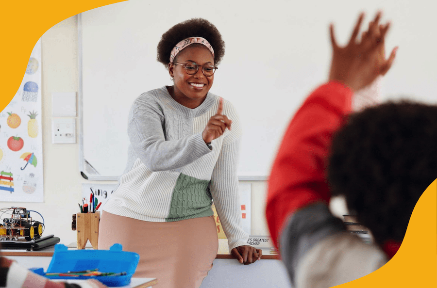 At the front of a classroom, a young woman holds up her index finger to call on the student raising his hand in the foreground.
