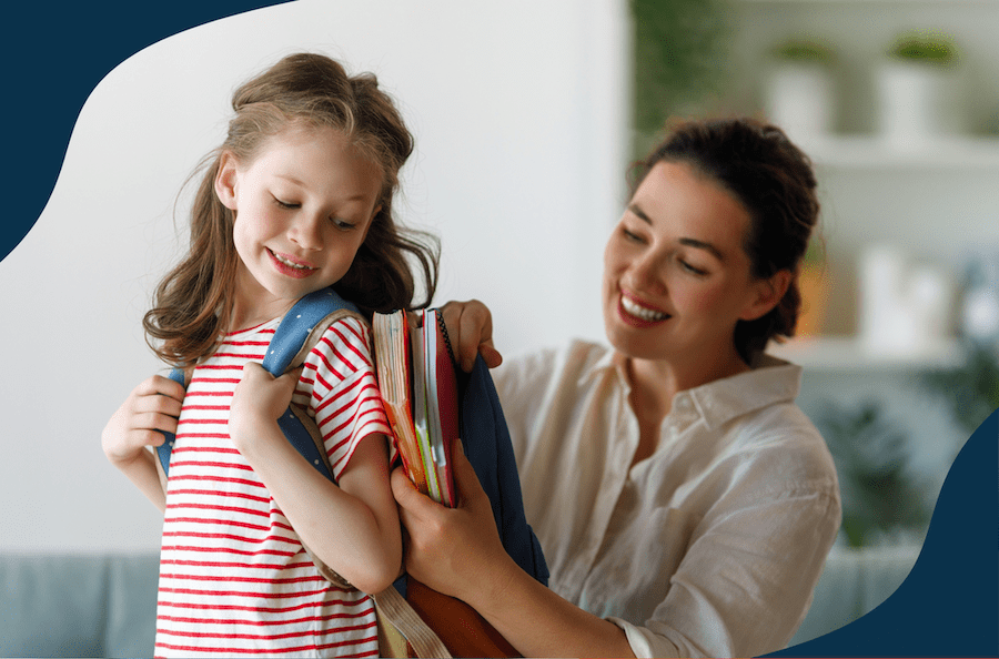A mother helps her daughter prepare for school. The young girl stands smiling with an open backpack while her mother puts books into the bag.