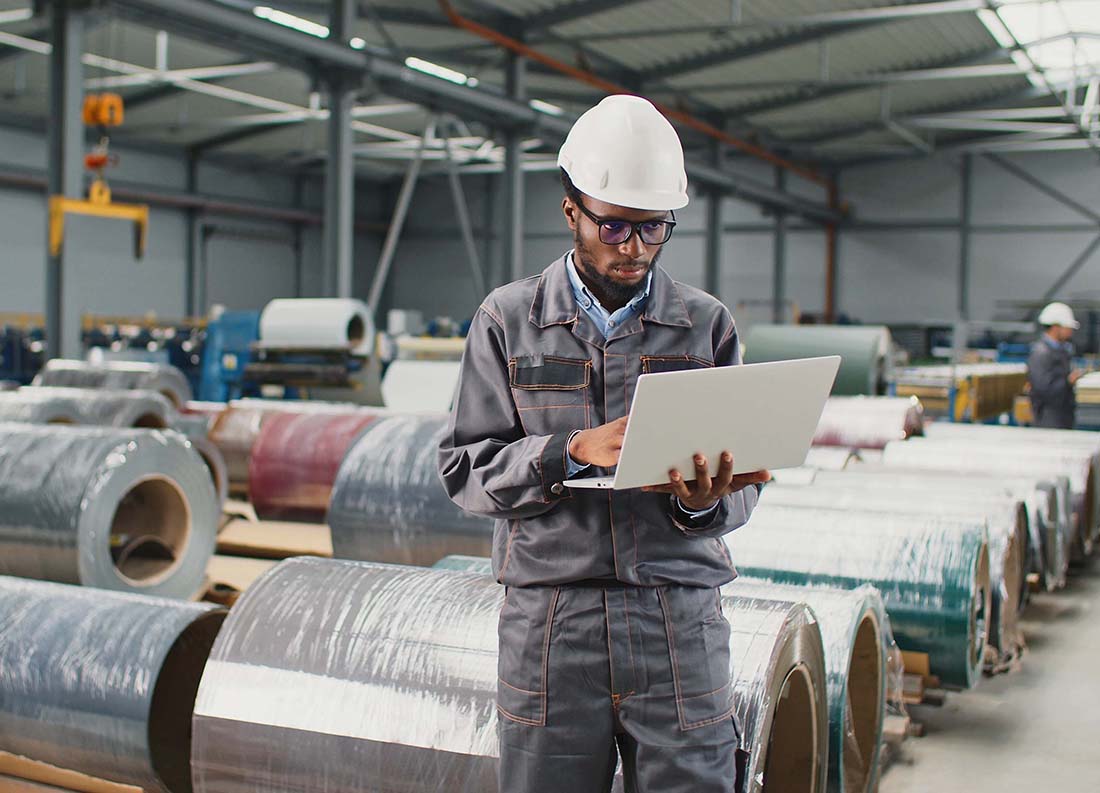 African American engineer using laptop at workplace. Worker wearing helmet and uniform standing in large storage warehouse.