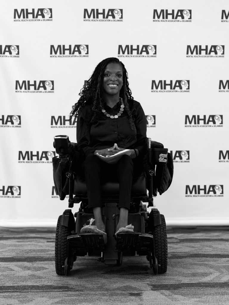 A woman sits in a wheelchair in front of background that says "MHA"